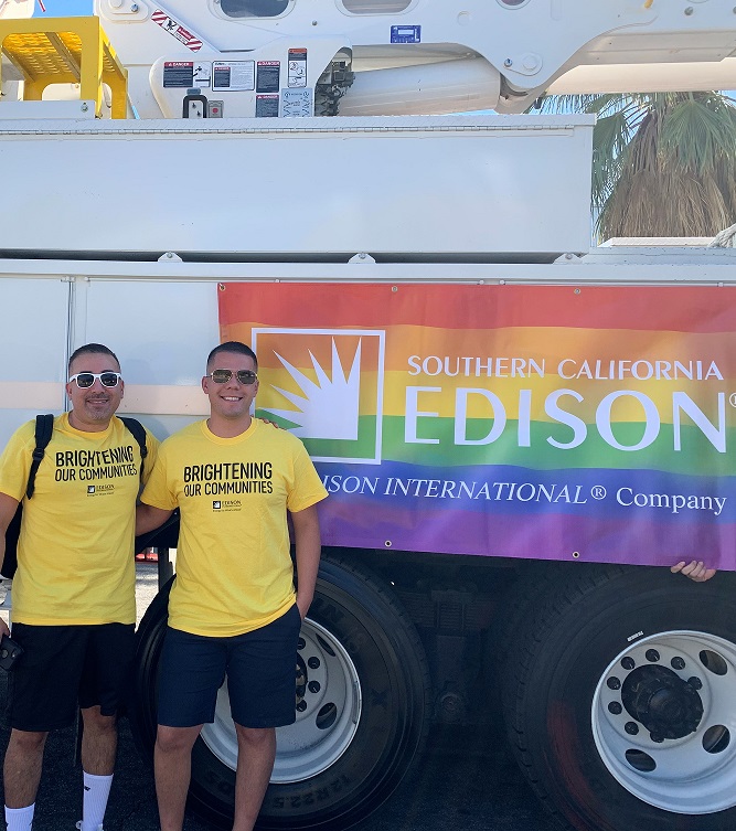 Two men smling and standing in front of a bucket truck with a sign behind them showing a rainbow flag and the company name: Souther California Edison. Edison International Company.