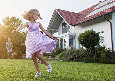 A girl with a purple dress playing in the front yard of a house and a woman in the background playing with a volleyball