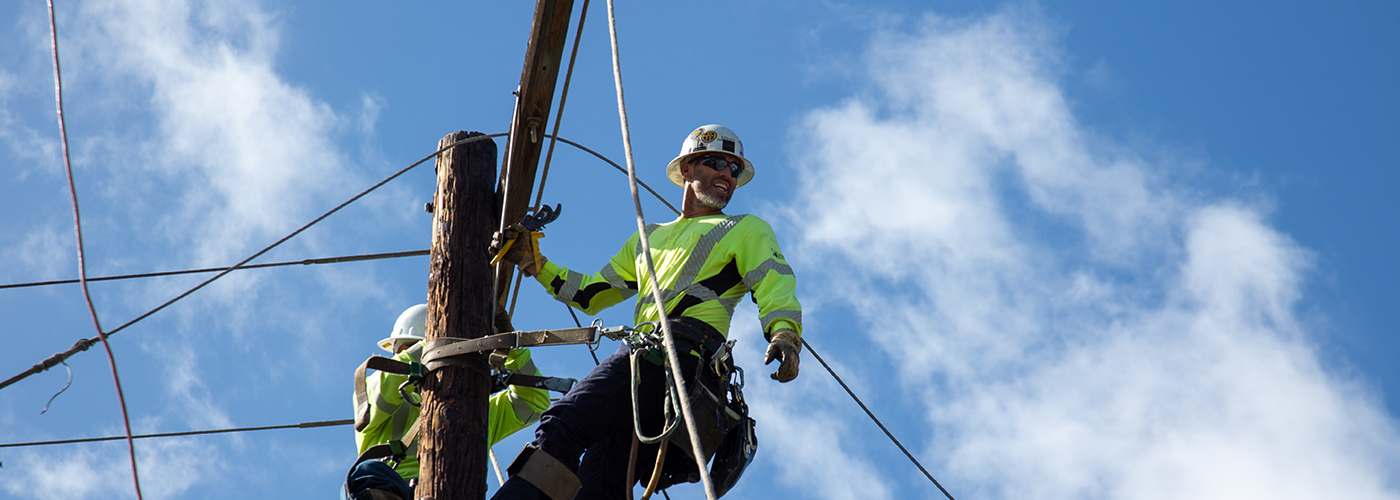 Two men on a pole wearing a hard hat and carrying tools looking away from the camera